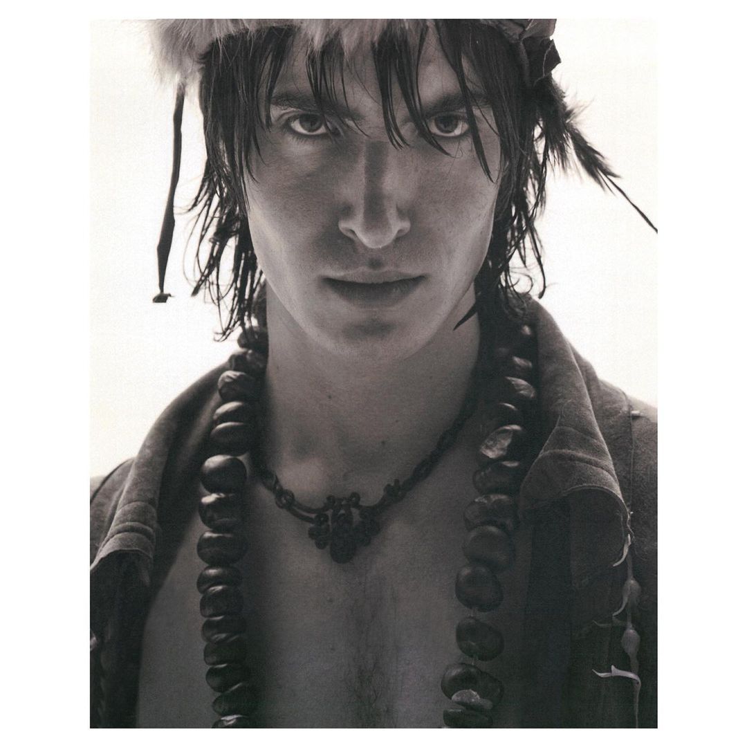 Monochrome portrait of a male model with dark hair wearing feathered headdress and beaded necklaces in a light studio setting.