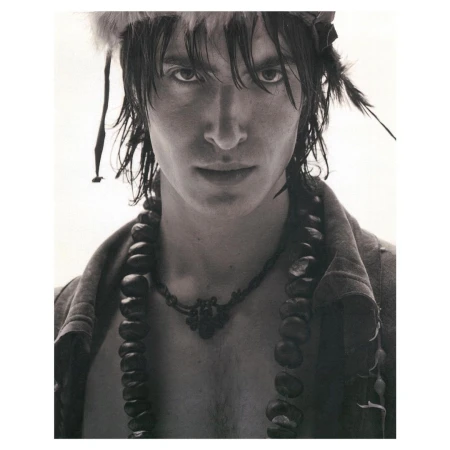 Monochrome portrait of a male model with dark hair wearing feathered headdress and beaded necklaces in a light studio setting.
