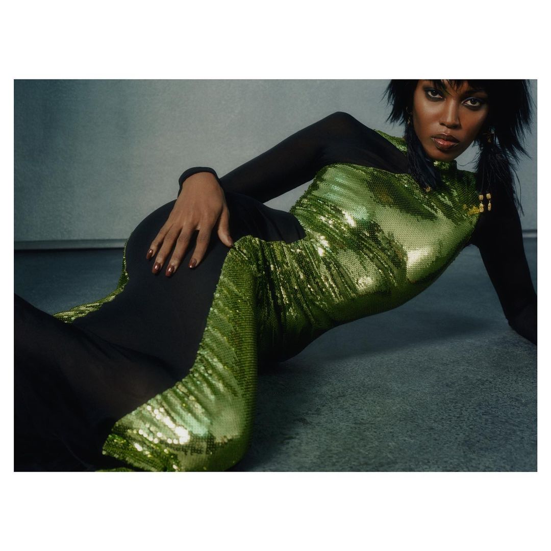 Woman with dark hair in a green sequin dress with black sheer sleeves, lying on a gray surface in a studio setting. Fashion shot.