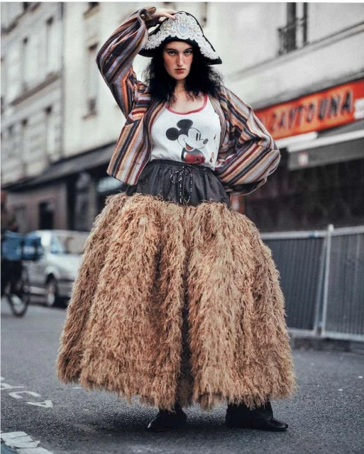 A woman wearing a large skirt and patterned jacket poses in an urban street setting, looking directly at the camera. Buildings in background.