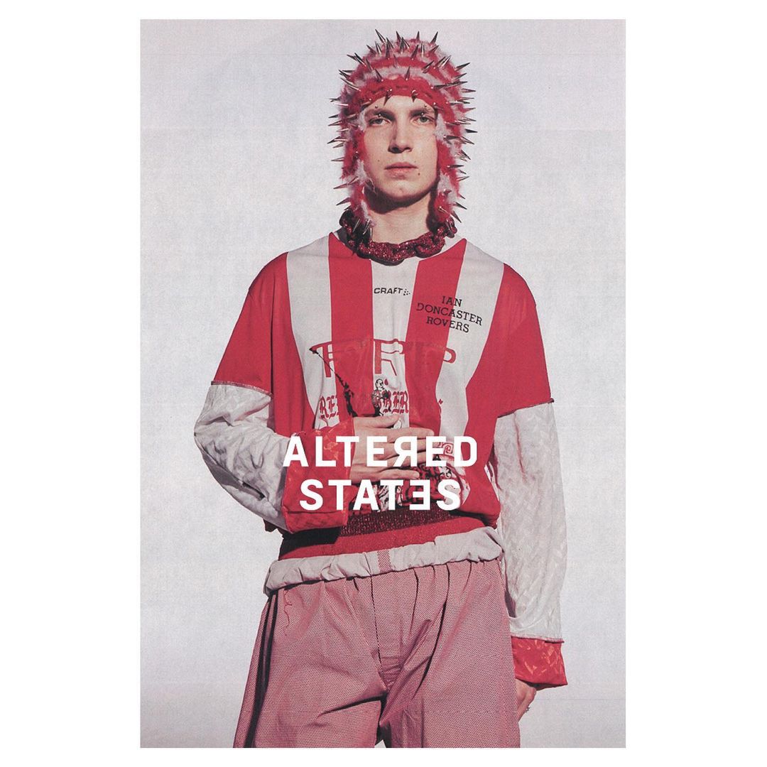 Fashion portrait of a fair-skinned male adult with spiked headwear, vertical striped top, and red patterned pants on a textured backdrop.
