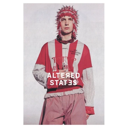 Fashion portrait of a fair-skinned male adult with spiked headwear, vertical striped top, and red patterned pants on a textured backdrop.