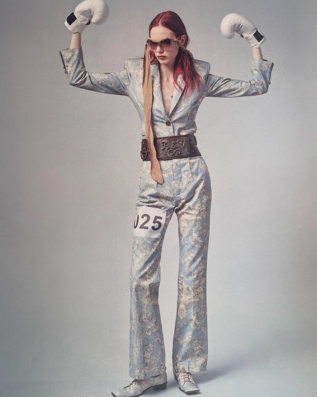 Woman in a patterned suit poses with boxing gloves in a studio setting. She is wearing sunglasses and has a number on her pants.