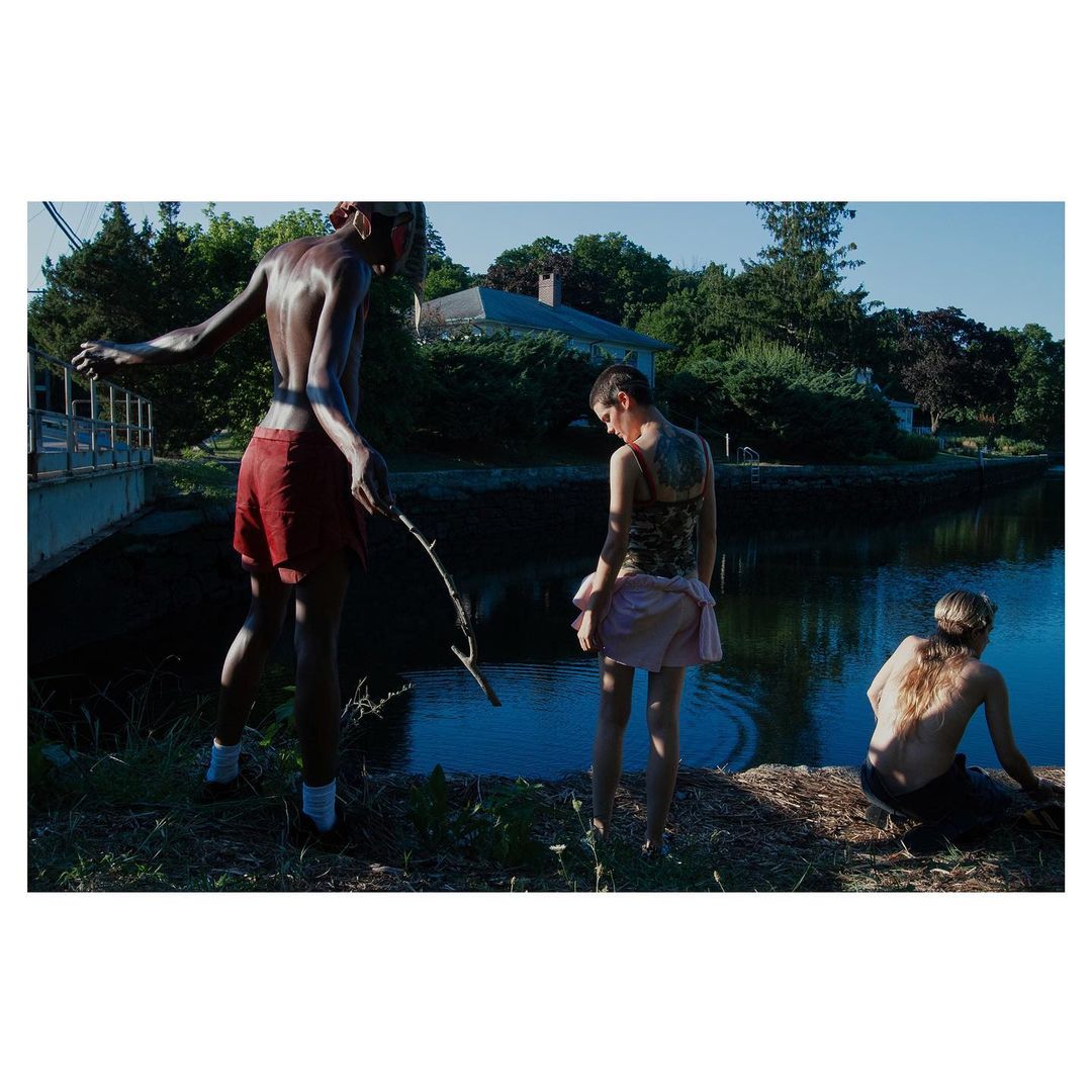 Three young adults relax by a canal in natural light. One figure holds a stick, creating a moment of tranquility and connection to nature.
