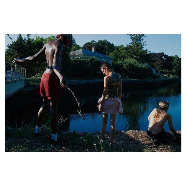 Three young adults relax by a canal in natural light. One figure holds a stick, creating a moment of tranquility and connection to nature.