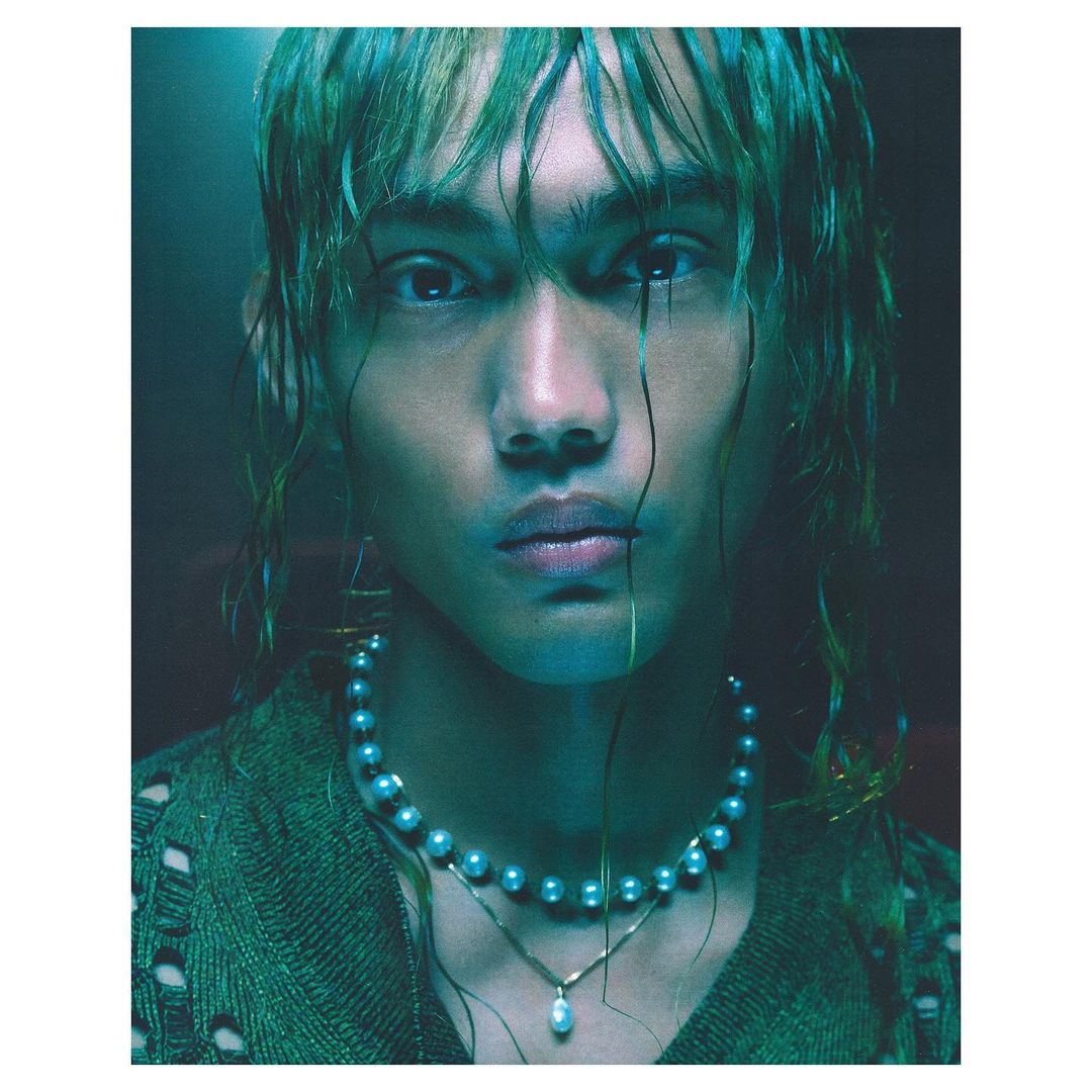 Portrait of a young adult with greenish dyed hair, wearing a pearl necklace and green knitwear, posing in a dark studio setting.