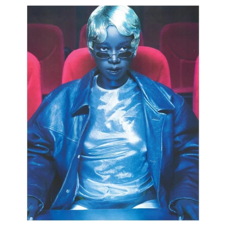 Young person with silvery hair and sunglasses wearing leather jacket and shiny top sitting in a red theater chair.