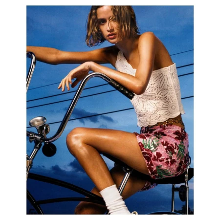 Woman in white patterned top and pink floral shorts sitting on a bicycle against a blue sky with power lines.