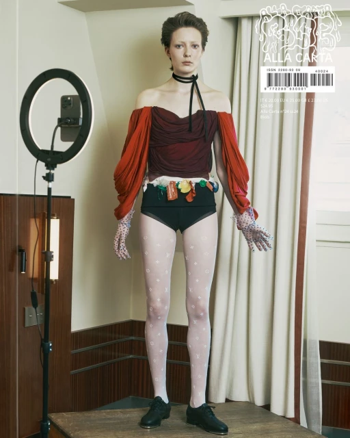Young woman in a haute couture fashion shoot wearing black shoes, sheer white patterned stockings, black high-waisted briefs, and a maroon top.