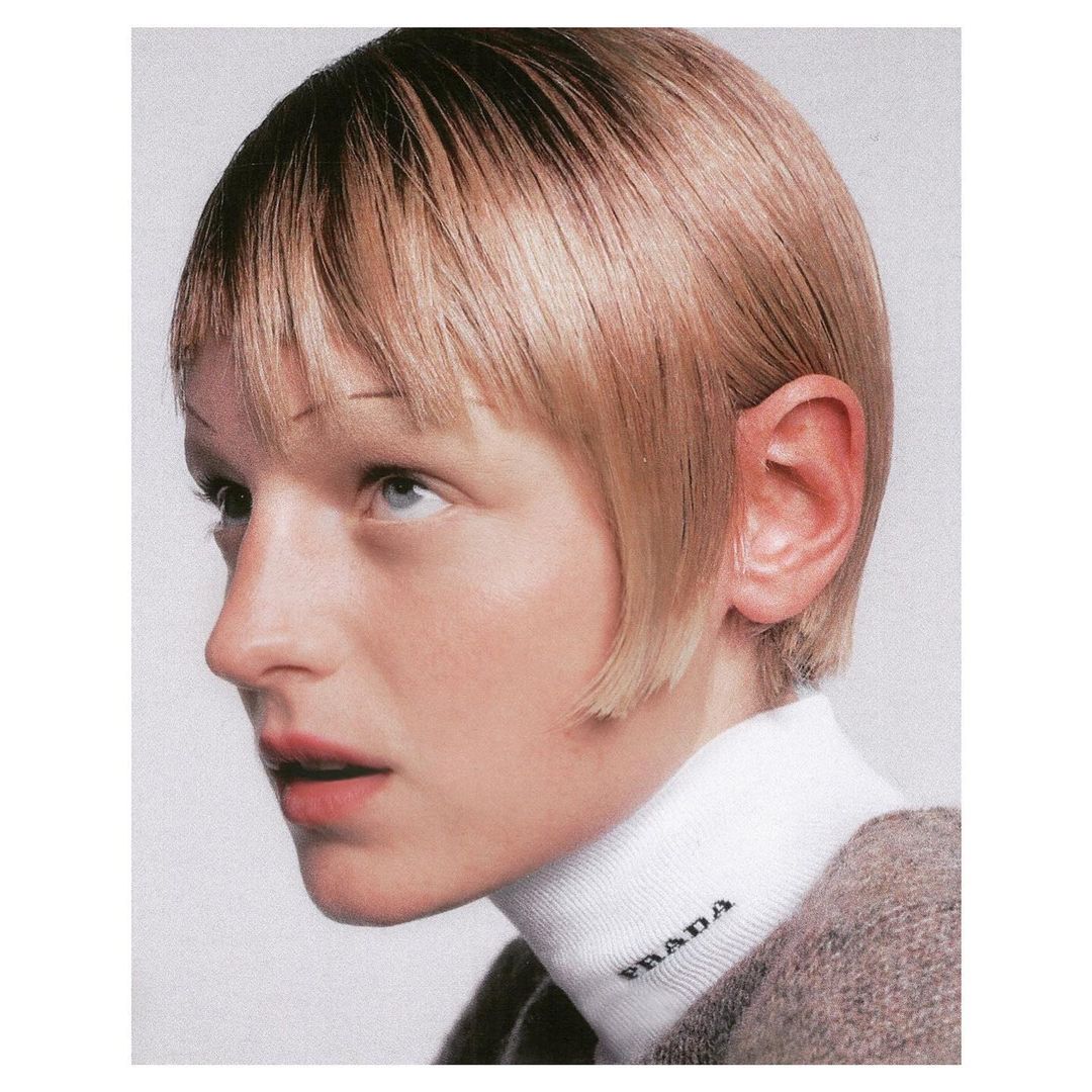 Close-up shot of an adult female with blond hair and fair skin wearing a white Prada turtleneck sweater in a bright, neutral setting.
