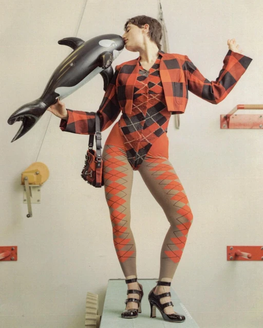 Fashion photo of a woman in an argyle bodysuit and plaid jacket, kissing a toy orca while standing on a platform indoors.