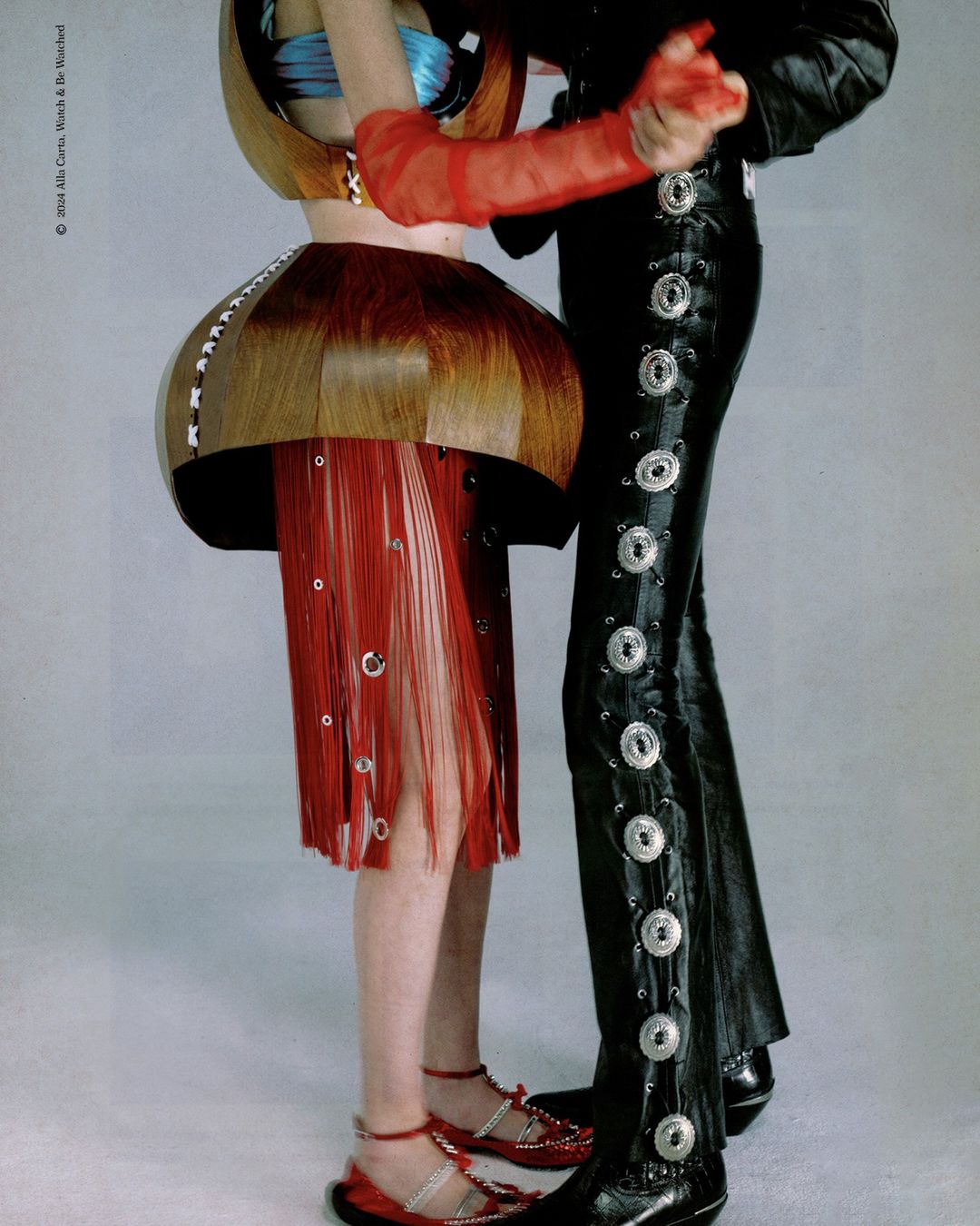 Two figures in avant-garde fashion, one in wood-like structured top and red fringe skirt, the other in embellished black pants and jacket.