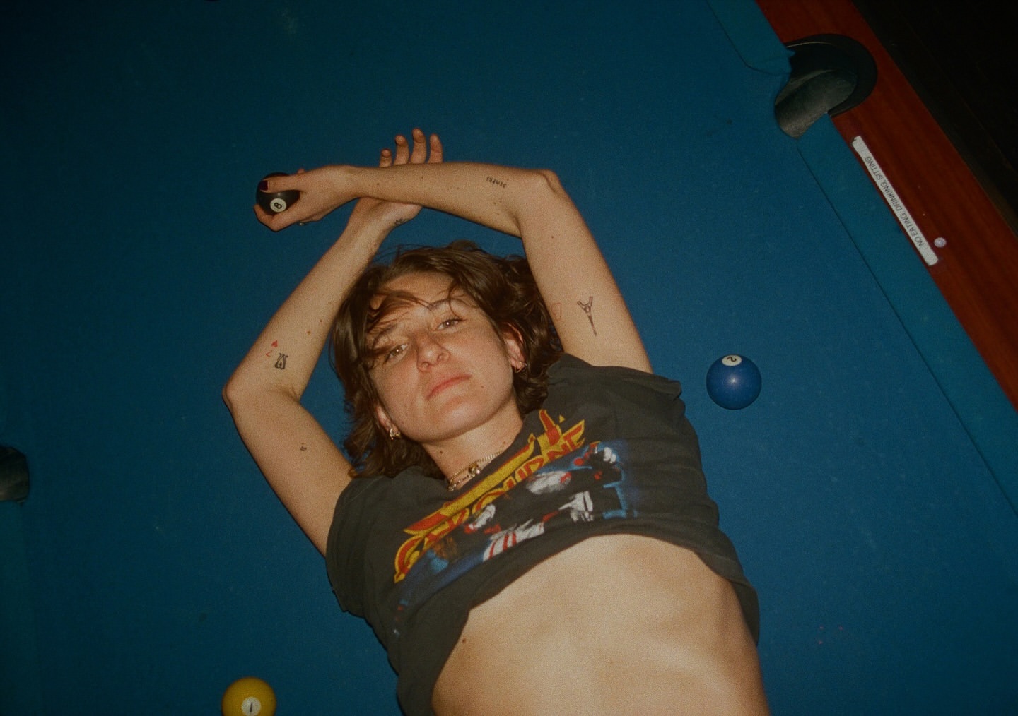 A young adult female lies on a billiard table with billiard balls, wearing a graphic tee. The scene is lit with indoor lighting.