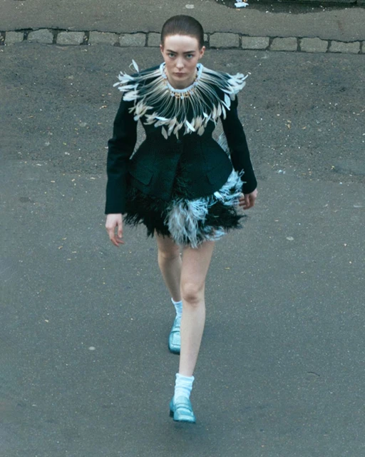 A woman in a black fashion jacket with feather trim is walking on an asphalt street and wearing light blue block-heeled shoes.