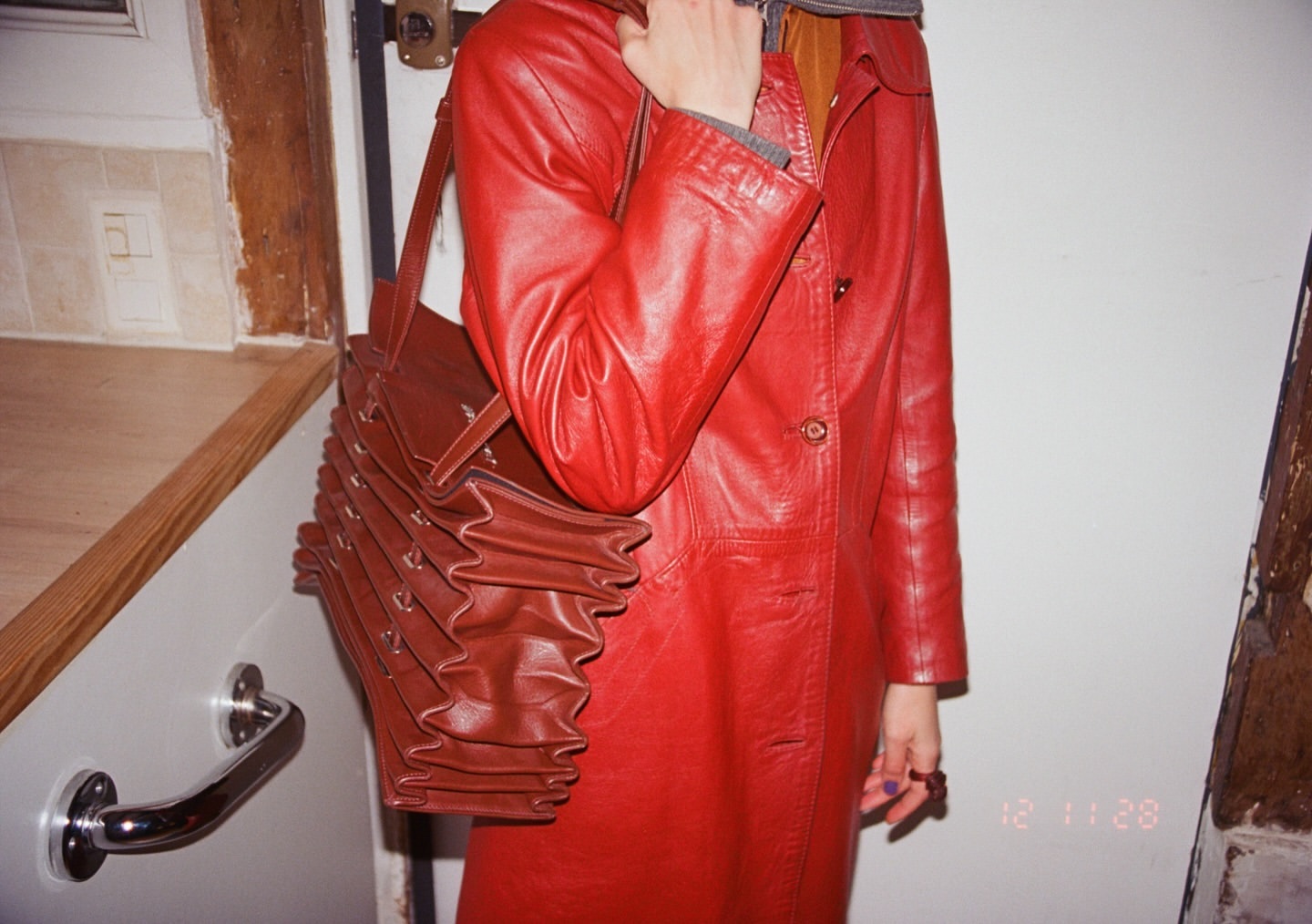 Red leather coat with an accordion purse. The garment is worn by an adult inside with white walls and a partial view of kitchen cabinetry.