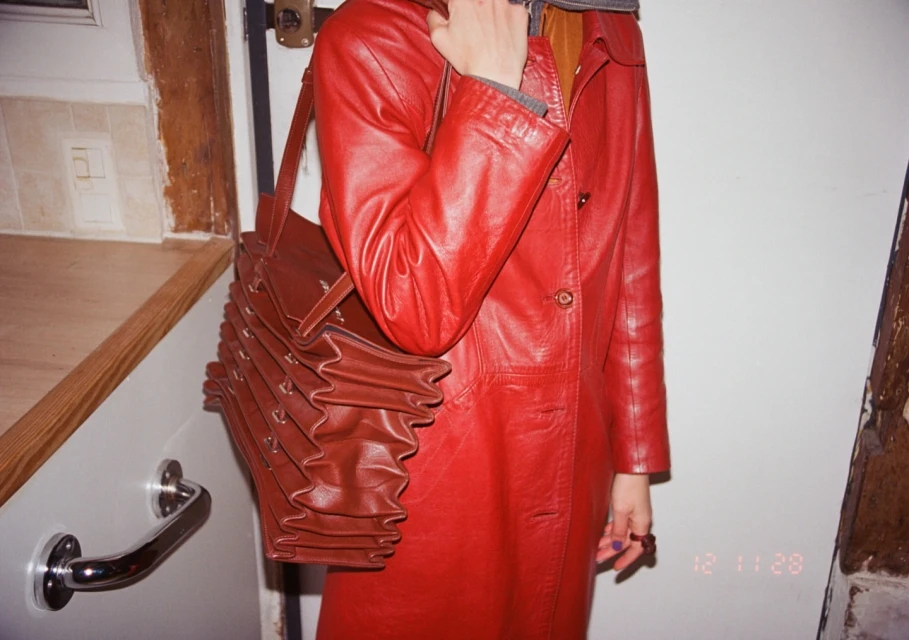 Red leather coat with an accordion purse. The garment is worn by an adult inside with white walls and a partial view of kitchen cabinetry.