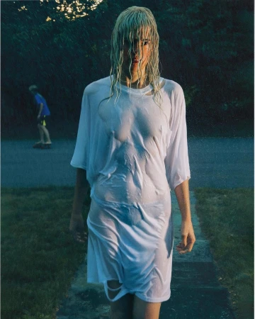 Woman with wet hair walking on stone path wearing wet white t-shirt dress in an outdoor environment with a child skateboarding in background.