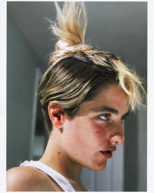 Close-up portrait of a young adult with blonde hair in a topknot, pale skin, wearing a white tank top, in an indoor setting.
