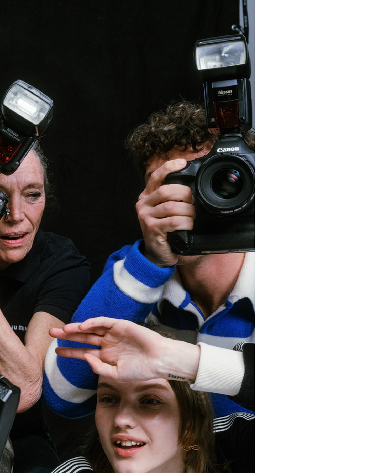 Three photographers capture a young woman in a photo studio. Two cameras with flashes are positioned around the woman's face. Black backdrop.