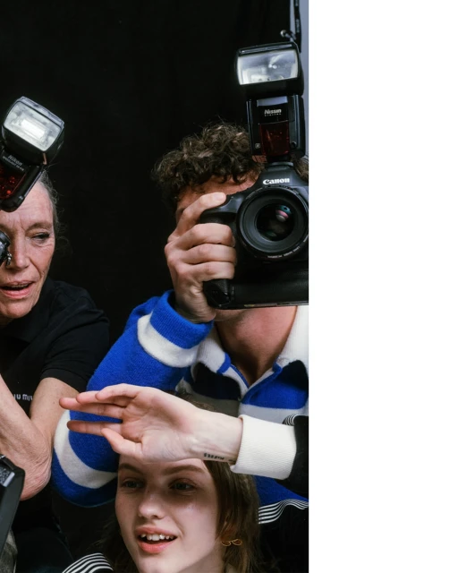 Three photographers capture a young woman in a photo studio. Two cameras with flashes are positioned around the woman's face. Black backdrop.
