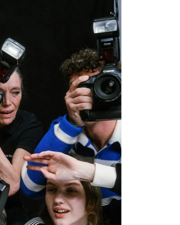Three photographers capture a young woman in a photo studio. Two cameras with flashes are positioned around the woman's face. Black backdrop.