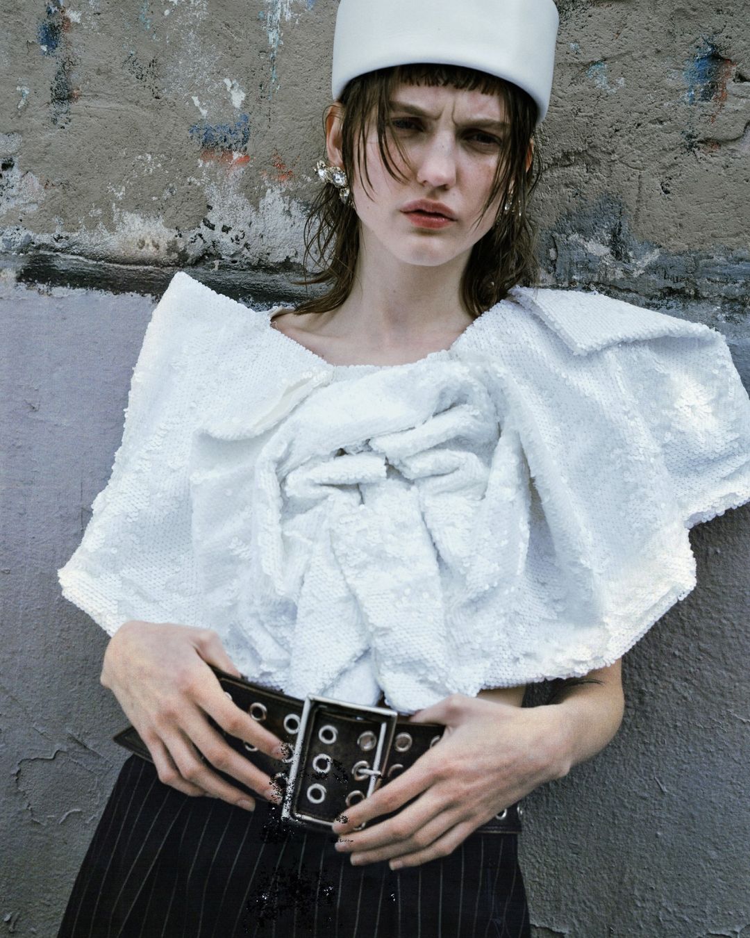 Fashionable young adult woman poses against a textured grey wall wearing a white sequin top, pinstripe pants and a white bucket hat.