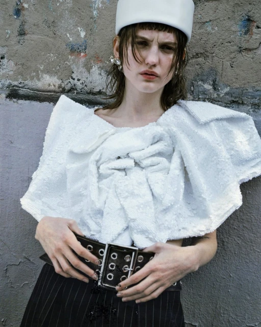 Fashionable young adult woman poses against a textured grey wall wearing a white sequin top, pinstripe pants and a white bucket hat.