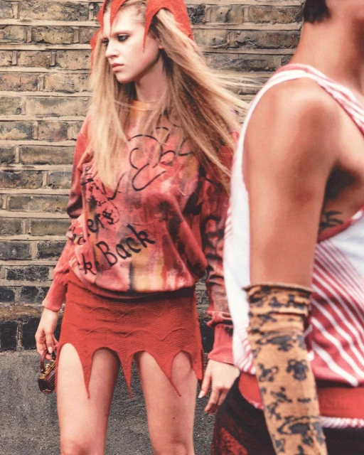 A woman in a red, edgy outfit with a patterned top and a man partially visible, posing against a brick wall, fashion editorial.