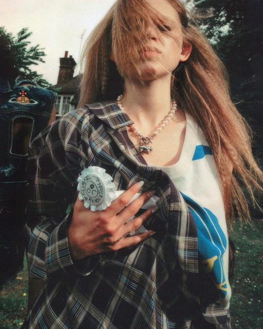 Young woman with crimped blonde hair, wearing a pearl necklace and plaid shirt poses outdoors in a close-up shot.
