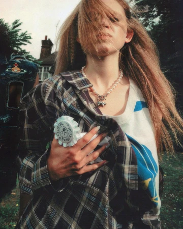 Young woman with crimped blonde hair, wearing a pearl necklace and plaid shirt poses outdoors in a close-up shot.