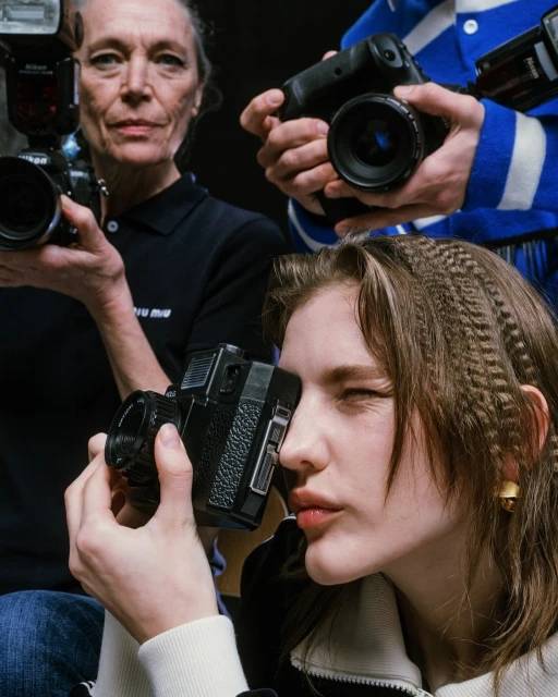 Three women holding cameras in a photo studio. A young woman in the foreground squints one eye and poses with a black camera.