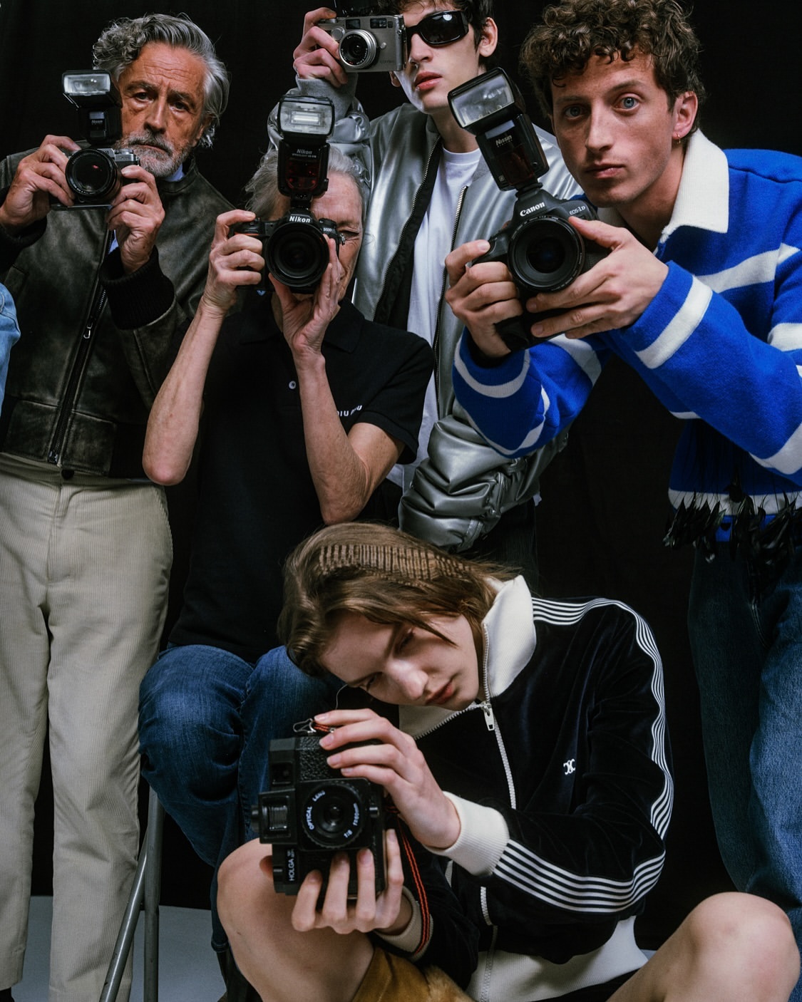 Group of people holding vintage cameras, some with flash attachments, in a photo studio with a black backdrop.
