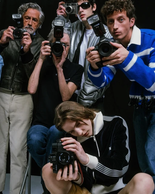 Group of people holding vintage cameras, some with flash attachments, in a photo studio with a black backdrop.