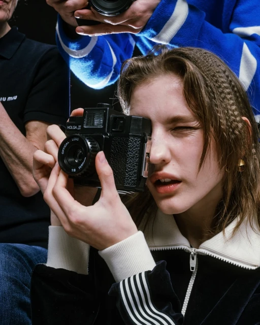 Young woman takes a photograph with a vintage camera, surrounded by other photographers in a studio setting with dark backdrop.