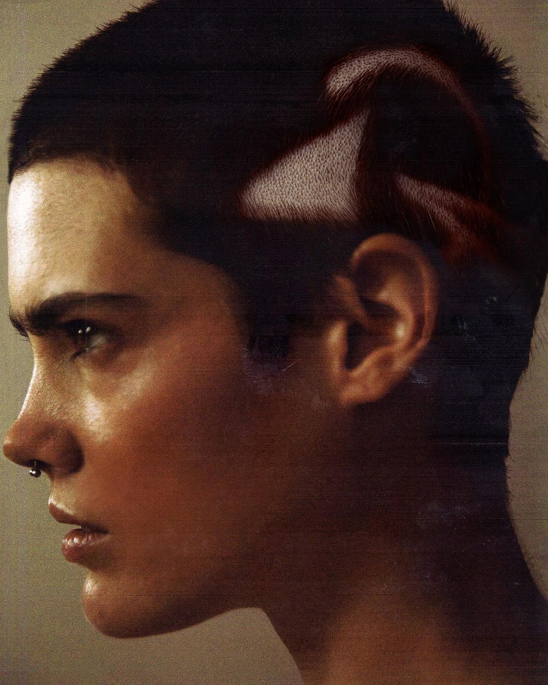 Close-up profile portrait of a person with short, dark hair and light skin. The person wears a nose ring and faces left.