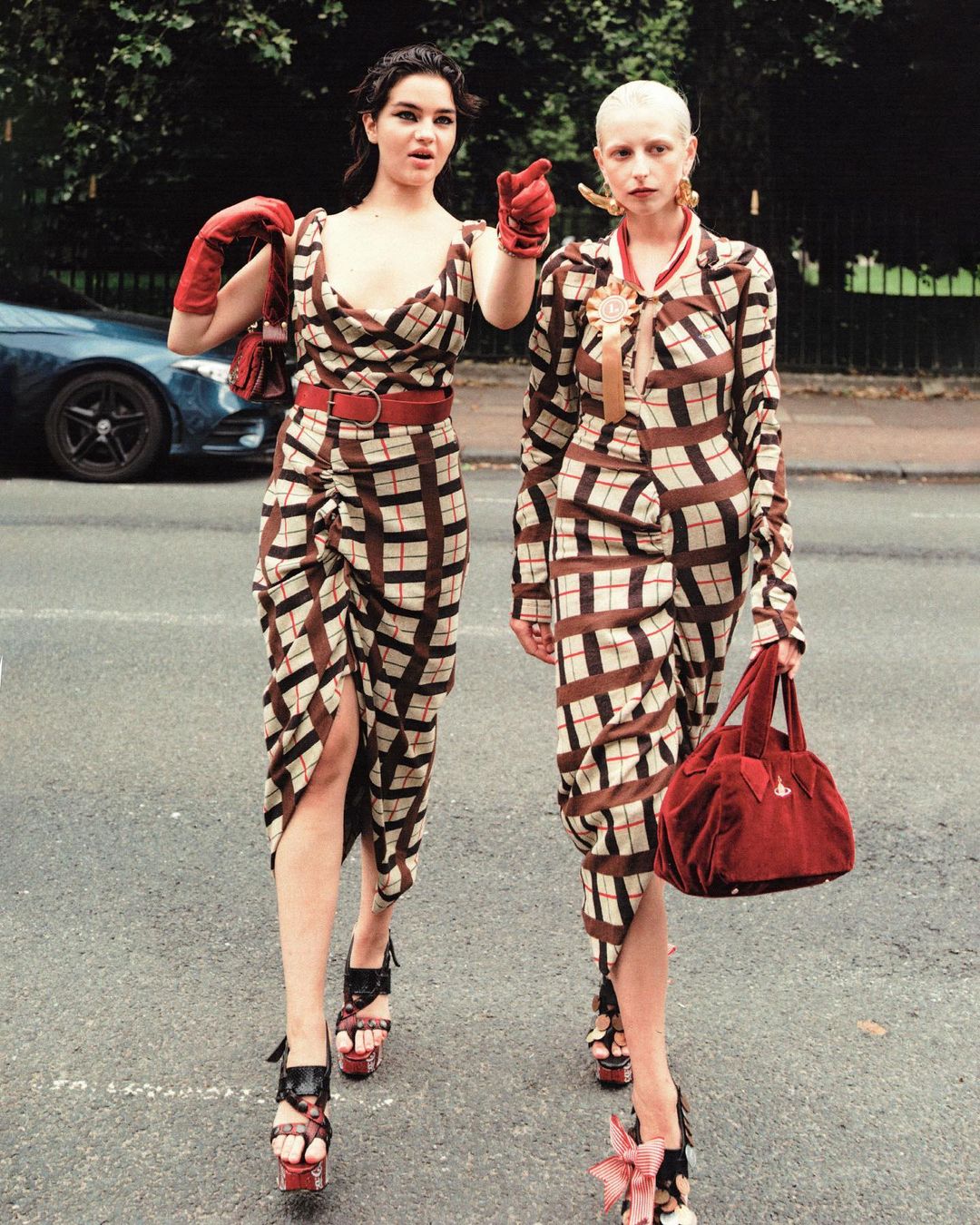 Two women in patterned dresses walk on a road outdoors, one pointing, both carrying bags and wearing platform heels.