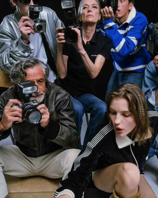 Several photographers of mixed ages, including a young model, are in a studio setting with dark backgrounds, showcasing cameras and fashion.