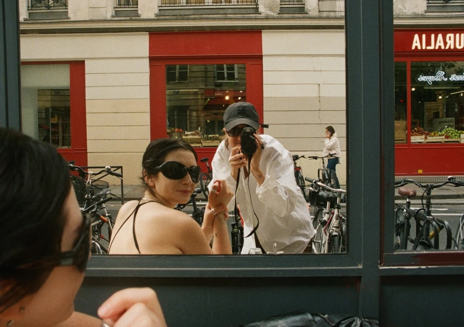 Two women in Paris are captured through a window: one with sunglasses looks toward the camera, while another takes a picture outdoors.