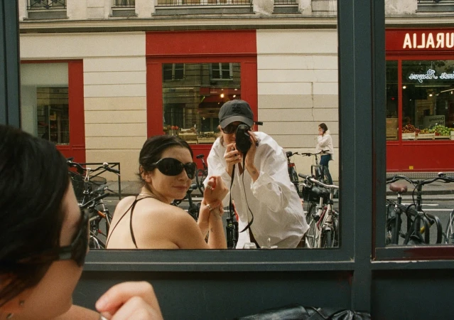 Two women in Paris are captured through a window: one with sunglasses looks toward the camera, while another takes a picture outdoors.