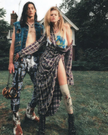 A fashion editorial presents a man and woman in alternative 90s-inspired outfits in a grassy, outdoor setting with a house behind them.