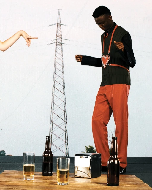 An adult male dances in an outdoor setting in front of an electrical transmission tower, with beer bottles and glasses on a wooden table.