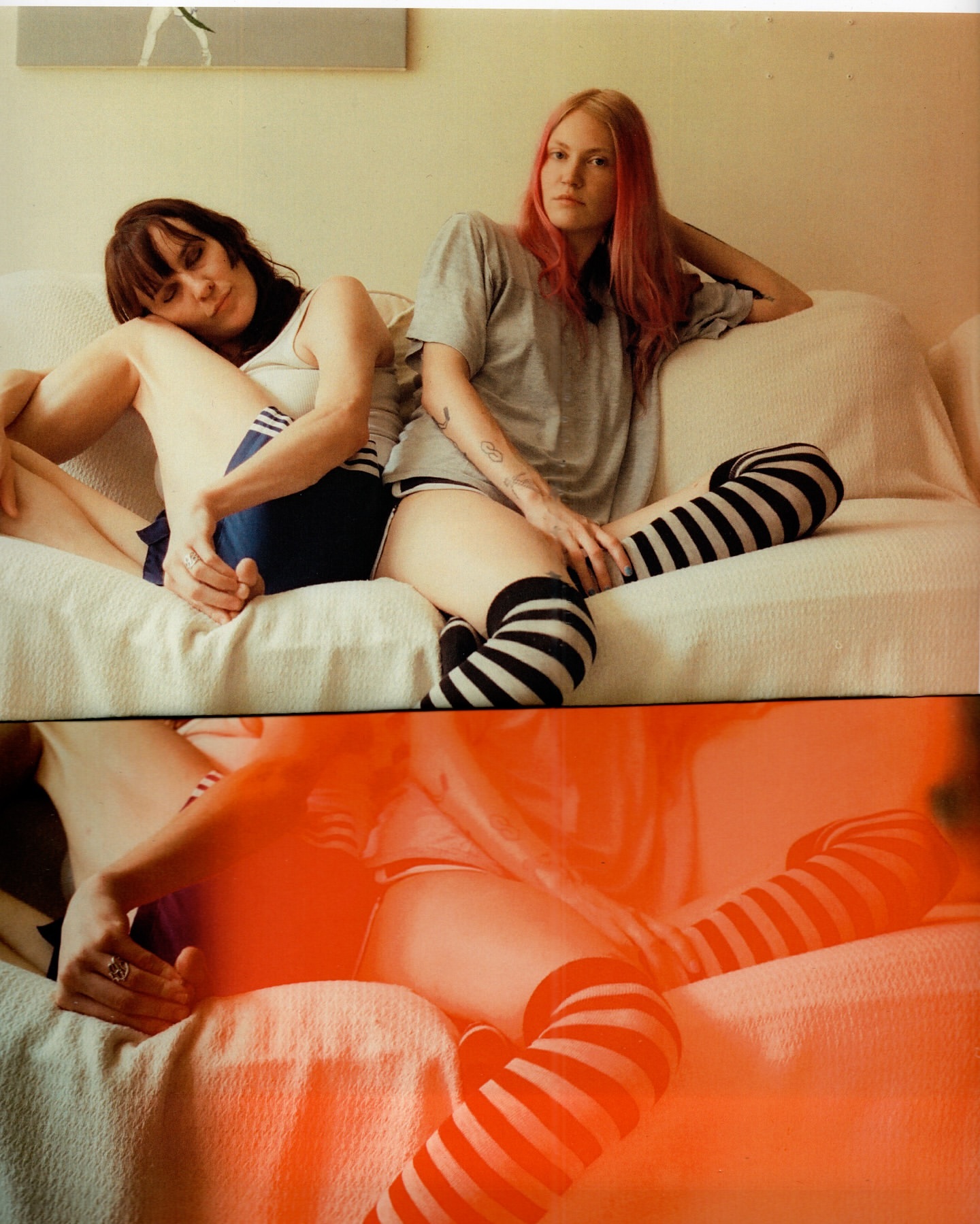 Two women lounge on a white sofa in casual clothing, one in athletic shorts and a tank top, the other in a t-shirt and striped socks.