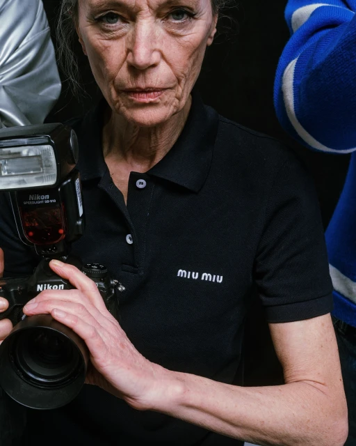 Woman in black polo shirt holds camera with flash in studio setting, surrounded by people partially visible, logo 'Miu Miu' visible.