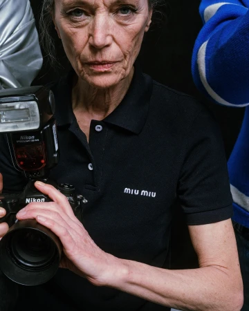 Woman in black polo shirt holds camera with flash in studio setting, surrounded by people partially visible, logo 'Miu Miu' visible.