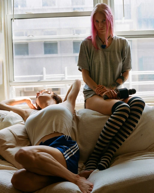Two young adult women lounge indoors near a bright window. One sits while holding an object, the other rests, wearing shorts and a tank top.
