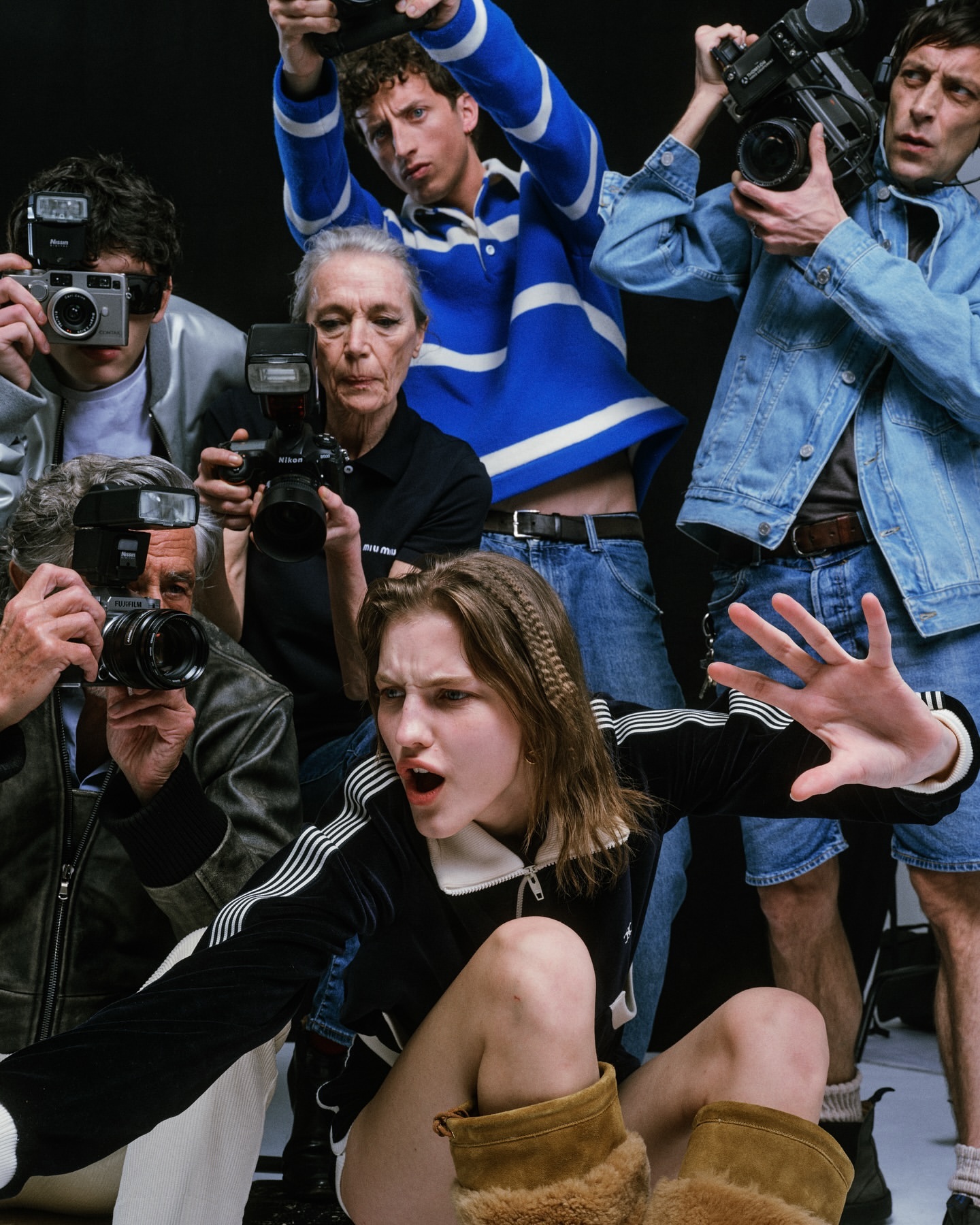 A group of paparazzi photographing a young woman in a black jacket and brown boots in a studio setting. The photographers are in jeans and jackets.