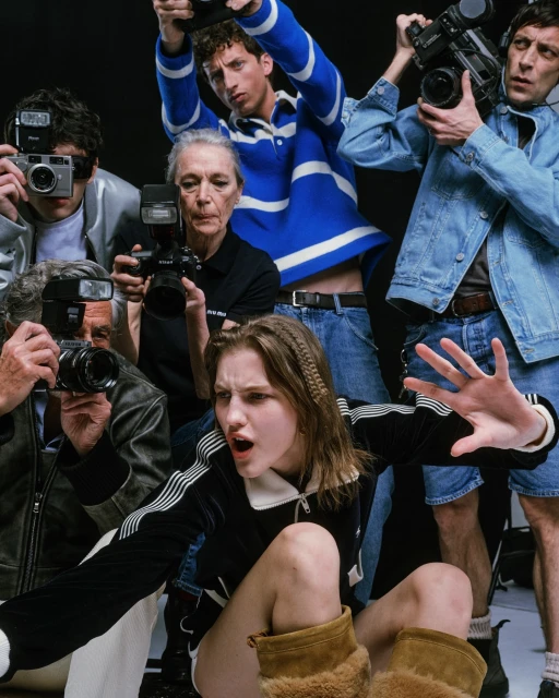 A group of paparazzi photographing a young woman in a black jacket and brown boots in a studio setting. The photographers are in jeans and jackets.
