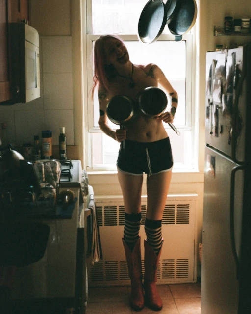 A woman in a kitchen holds up pots; light shines through the window behind her. She wears shorts, striped socks, and red cowboy boots.