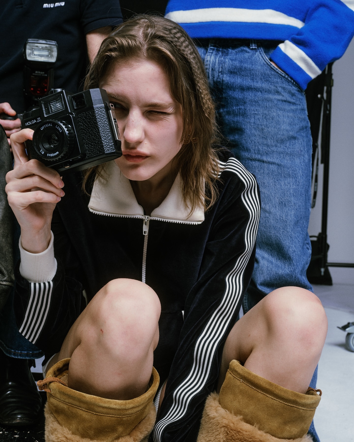 Young adult poses in a studio with a vintage camera, wearing a black velour tracksuit with white stripes and light brown UGG boots.