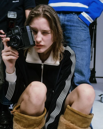 Young adult poses in a studio with a vintage camera, wearing a black velour tracksuit with white stripes and light brown UGG boots.